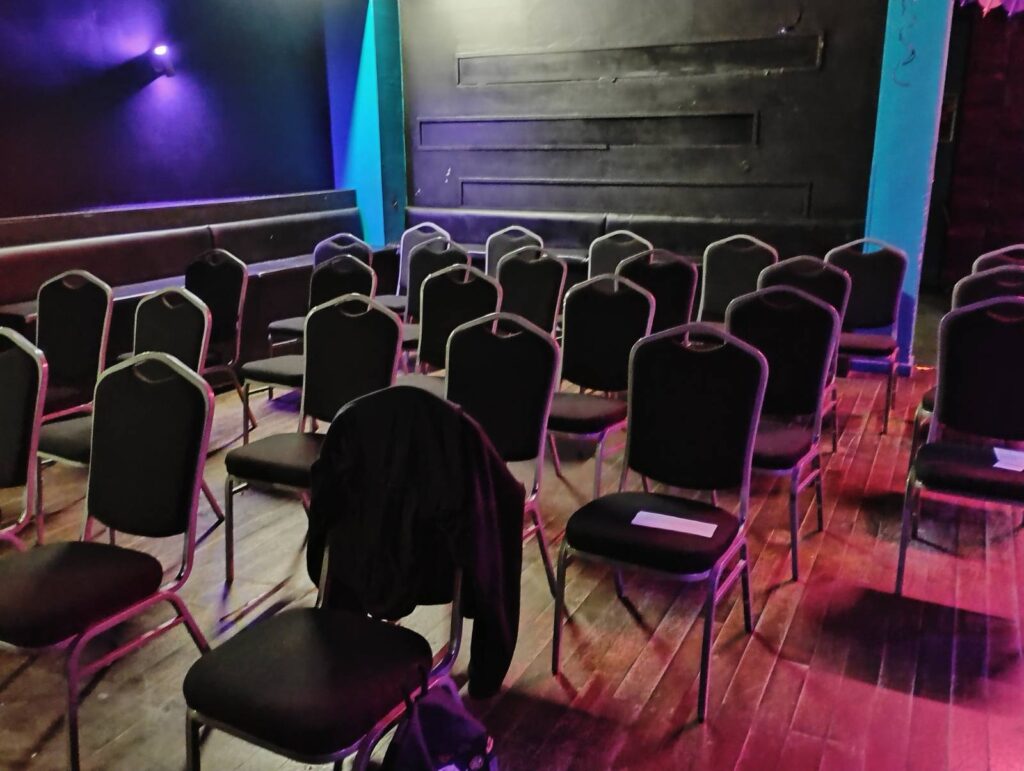a smallish room with a wooden floor and black panelled walls filled with black chairs, some which have handouts on them.