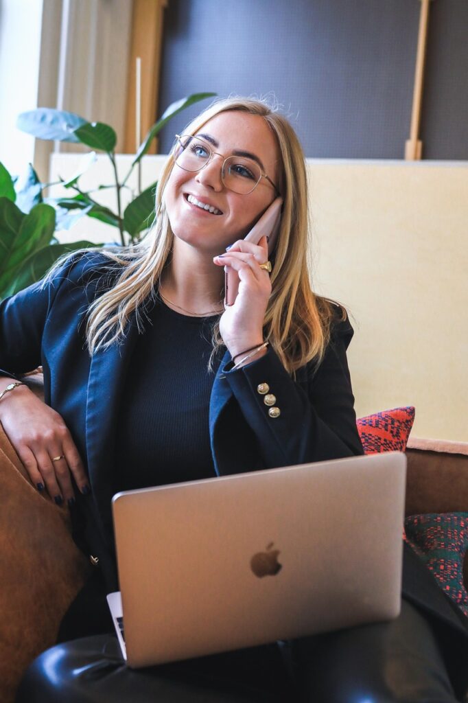 a woman with light skin tone and long blonde hair wearing a navy blue casual suit. She is sitting on a brown sofa with a mobile phone to her ear, a Macbook on her lap, and smiling.