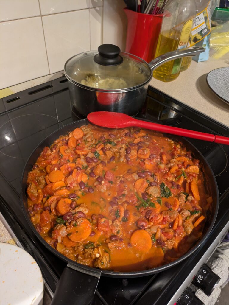 A large pan with a red sauce with carrots and other goodies in it cooking in it, another pan on the hob nearby has pasta cooking in it.