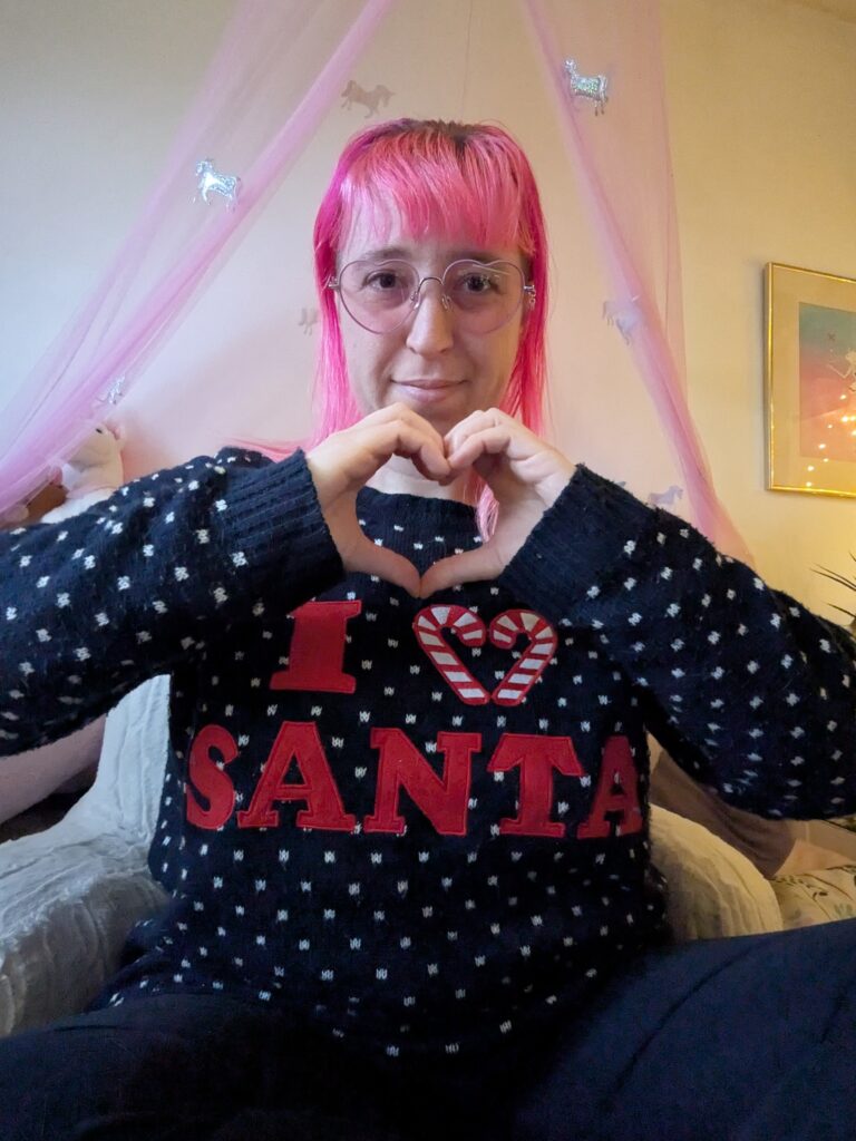 Alexis, a woman with light skin tone and bright pink hair, wearing pink tinted heart shaped glasses and a dark blue jumper with I heart Santa on it. She is sat on a bed making a heart shape with her hands and smiling.