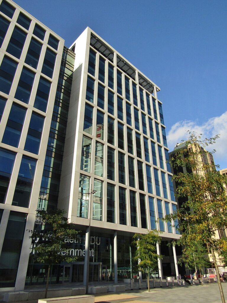 a large multistorey building with large windows, some trees barely 2 storeys in height are dotted around the mostly cement outside area.