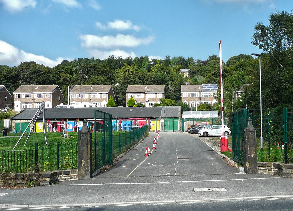 A road past a school field on the left surrounded by green fence with cones alongside it. At the far end of the road are garages with different coloured doors and some cars parked beside them. Behind those are some large houses and a wooded area. The sky above is blue.