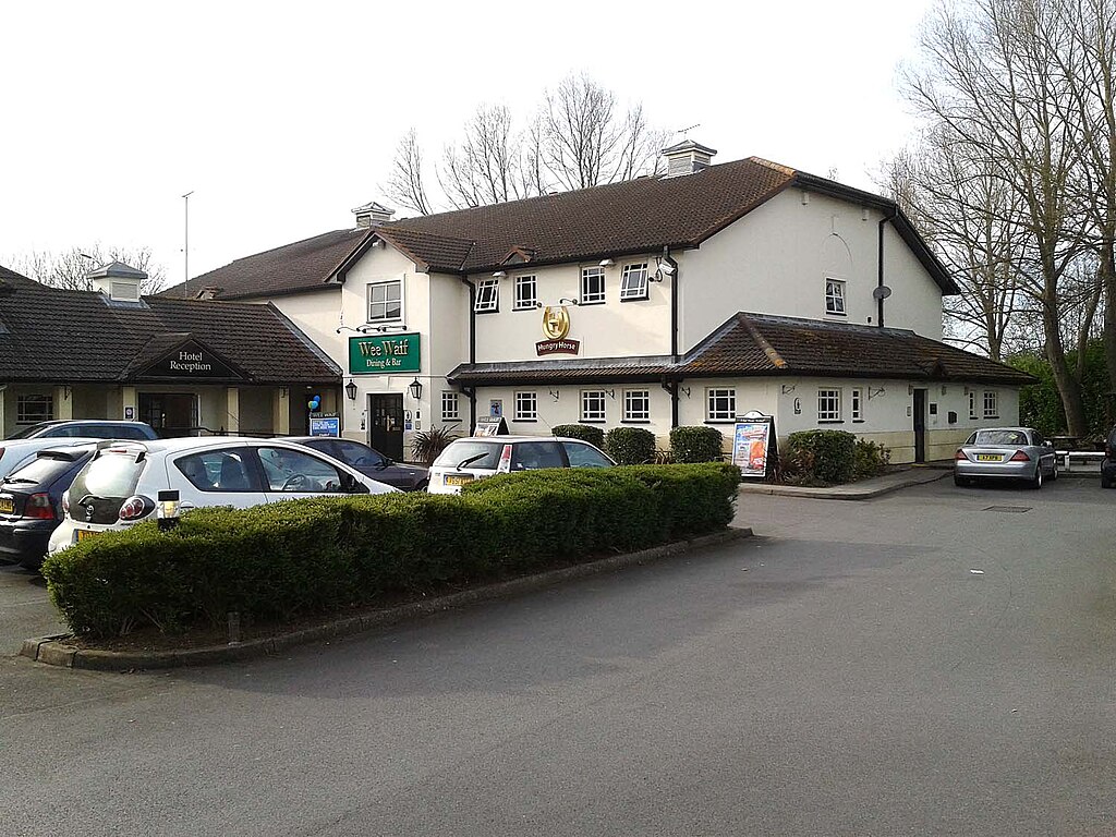 a British pub, a large beige building with tiled roof and a sign above the door reading “Wee Waif.” In front of the building is a car park filled with cars.