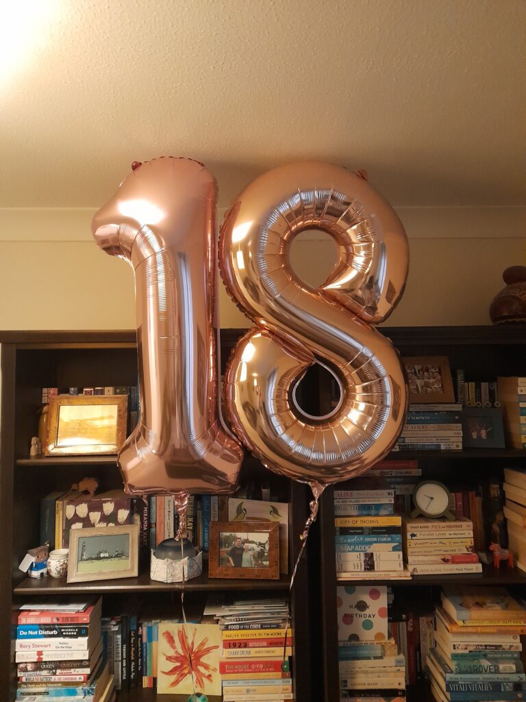 a large, rose gold balloon of the number 18 floating in front of a very full bookcase stacked with books and photos in frames.