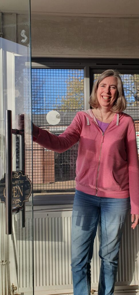 Sarah, a woman with light skin tone and shoulder length light brown hair, wearing a pink hoodie and jeans. She is standing holding a door open and smiling broadly as the sun shines through the window behind her.