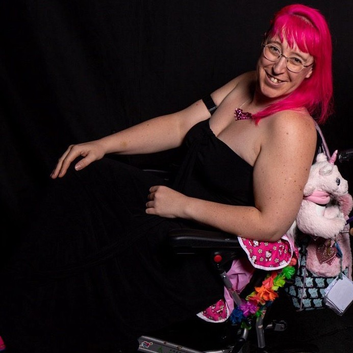 a woman with light skin tone and long bright pink hair wearing a black evening dress and sitting in an electric wheelchair, smiling broadly. On the back of the chair is a unicorn and a pink fluffy bag.