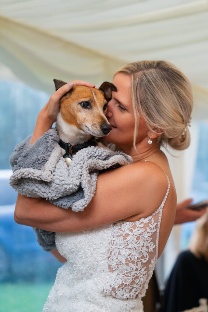 Lisa, a woman with light skin tone and blonde hair in a beautiful up-do. She is wearing a wedding dress and holding tight an elderly Jack Russell wrapped in a fluffy blanket.
