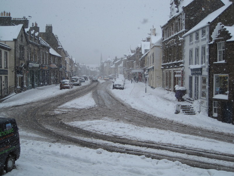 A snowy road through a Scottish street, cars are parked either side covered in snow. The grey sky and falling snow make it look especially cold.