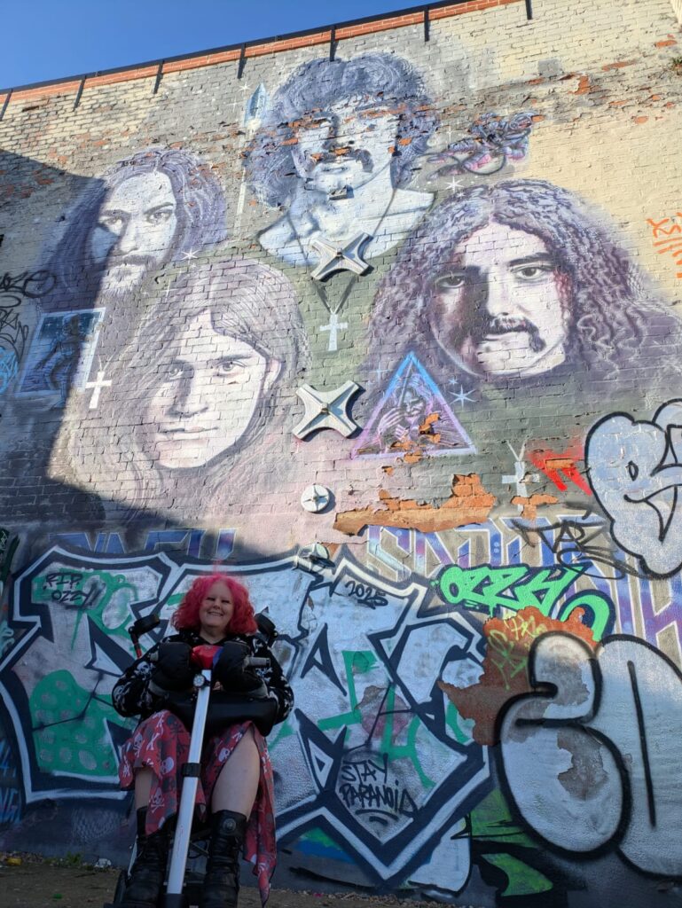 a woman with light skin tone and long curly red hair wearing a black jacket and red handkerchief skirt. She is sitting on a mobility scooter under a mural of Black Sabbath on a wall