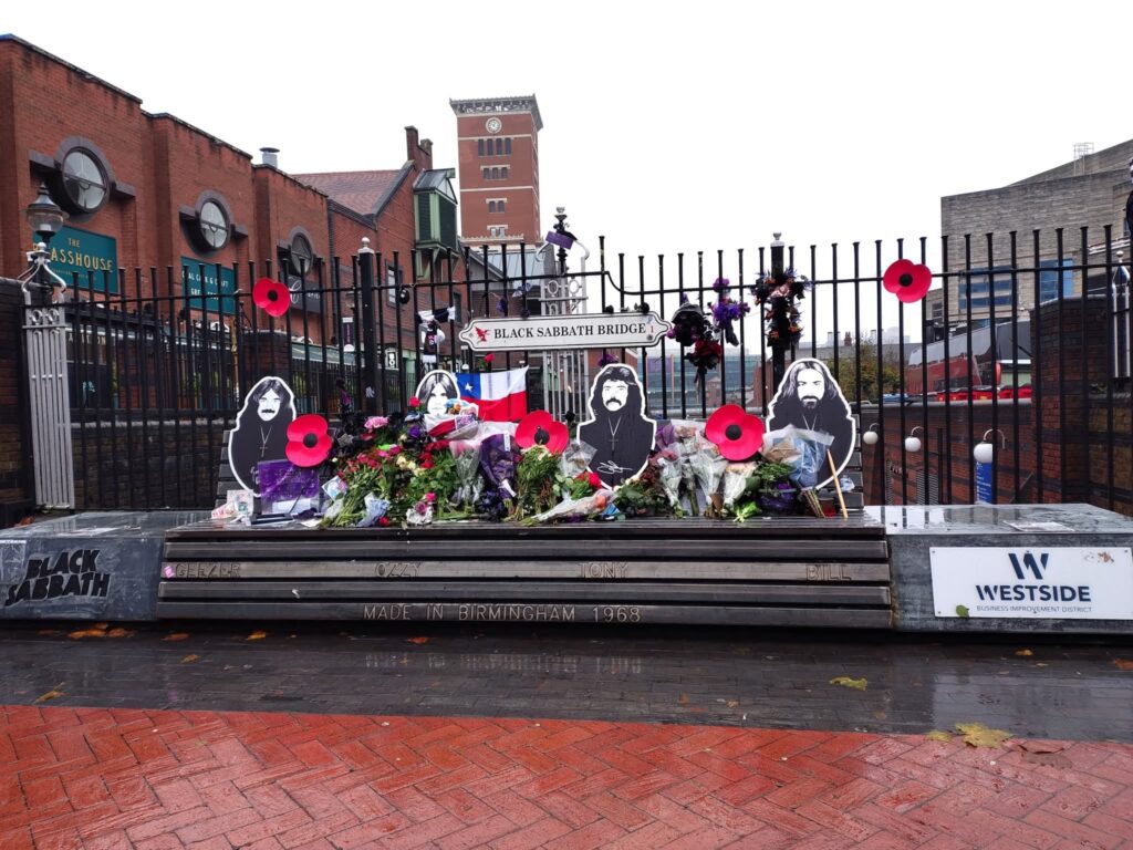 the Black Sabbath bench in Birmingham, a bench with cut outs of the band members attached to it. There are also poppies and flowers laid on the bench. It looks like a very wet and dreary day.