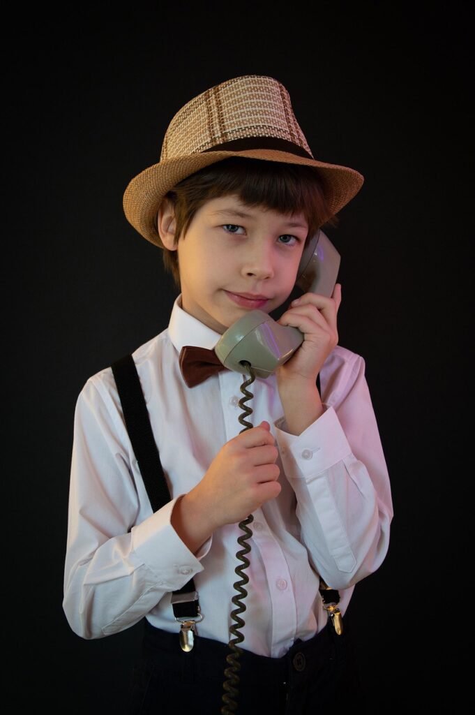 a young boy dressed in a shirt, bow tie, suspenders and a trilby hat holding an old fashioned corded phone to his ear.