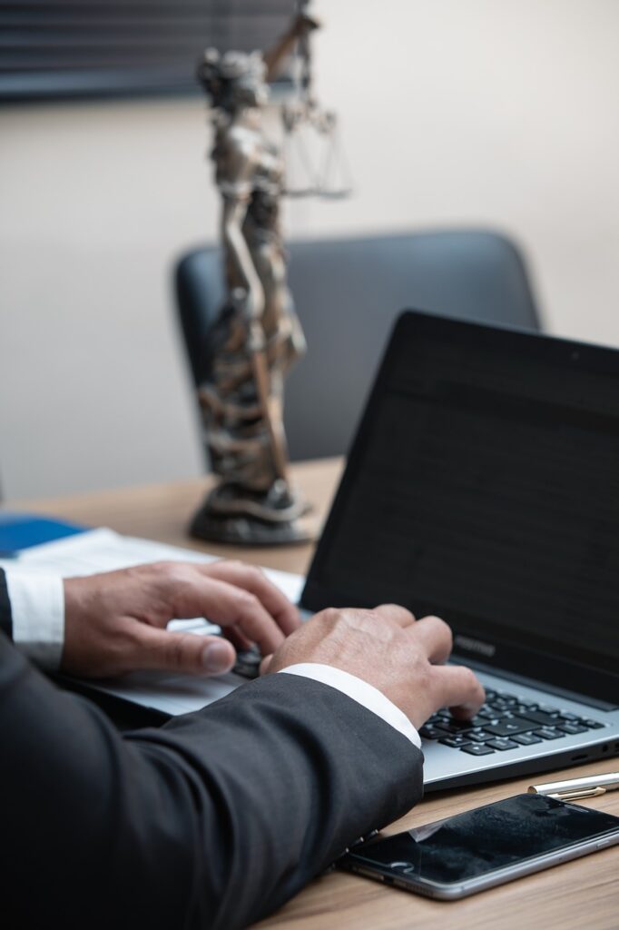 hands coming from a suit typing on a laptop with a phone beside them. In the background is a small sculpture of Justice holding scales sat on the desk indicating it is the office of a lawyer.