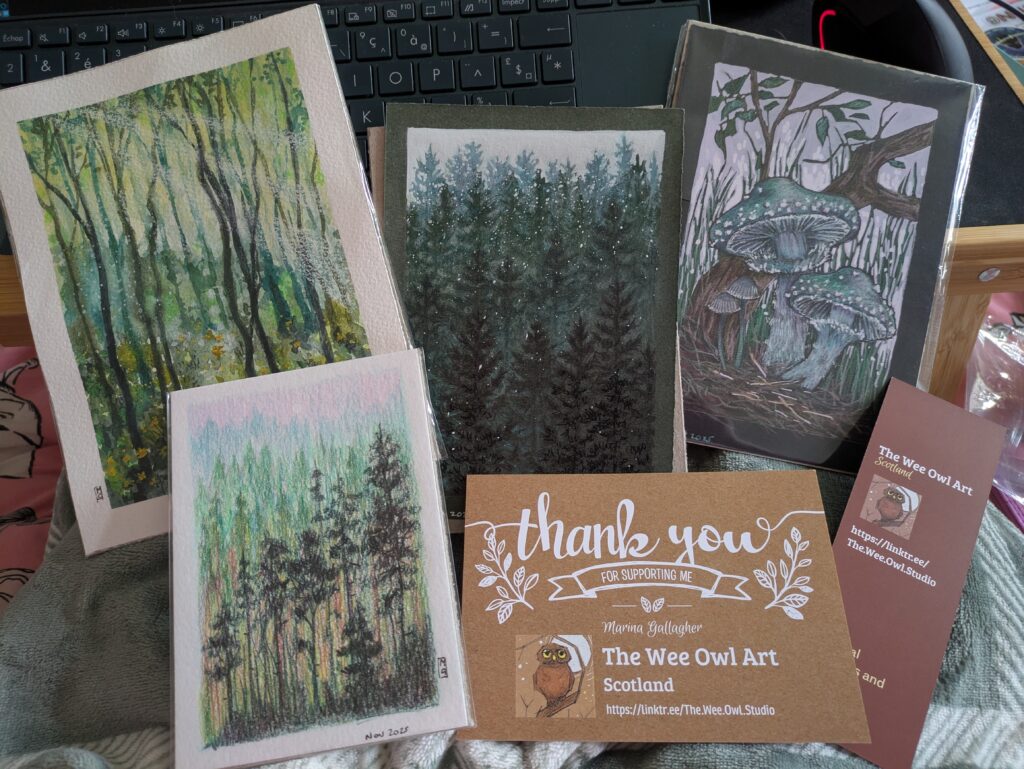 Four artworks resting on a bed beside a thank you postcard from The Wee Owl Art and a bookmark from them. The first is a spring forest with tall slim trees and light streaming in. The second a dark wintery forest with snow falling on it. The third green mushrooms growing out of the soil by a tree branch, they are huge. The fourth is a smaller lush green forest with some autumn colours in it. All heavily feature different colours of green.