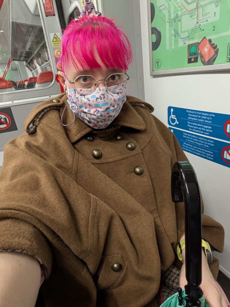 a woman with light skin tone and bright pink hair wearing glasses, a pink face mask with unicorns on it, and a brown cape with bronze buttons while holding an umbrella, sitting in a wheelchair on a train.