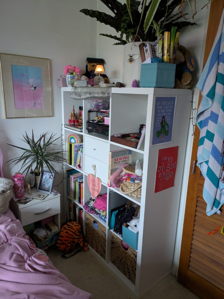 a 3 by 4 cube storage unit filled with two baskets at the bottom, books in three cubes, folded clothes in one, two drawers in one, knick knacks and sewing bits in others. A plant and decoration on the top.