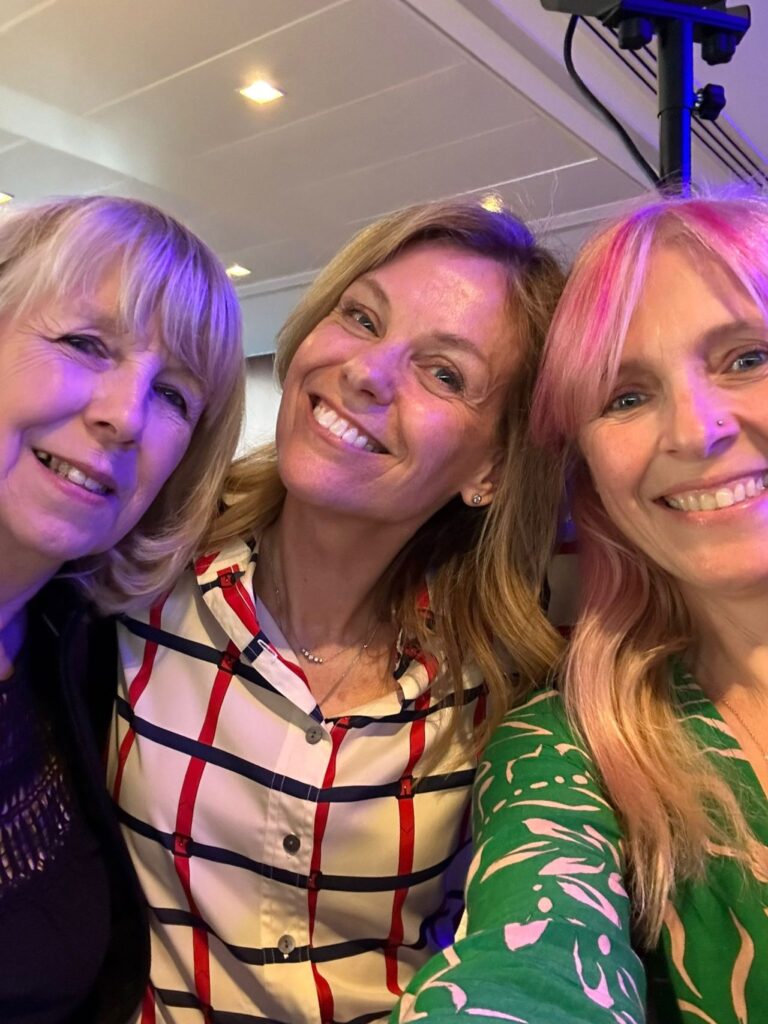 three women of different ages, all with light skin tone and long blonde or light brown hair. They are all leaning into each other and smiling broadly.