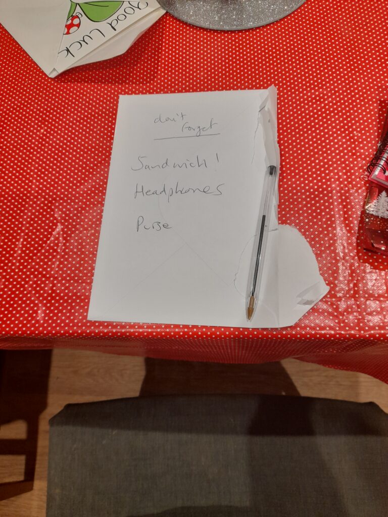 an opened envelope with don’t forget, sandwiches, headphones, purse written on it and a pen resting on it. The envelope rests on a red tablecloth on a table.