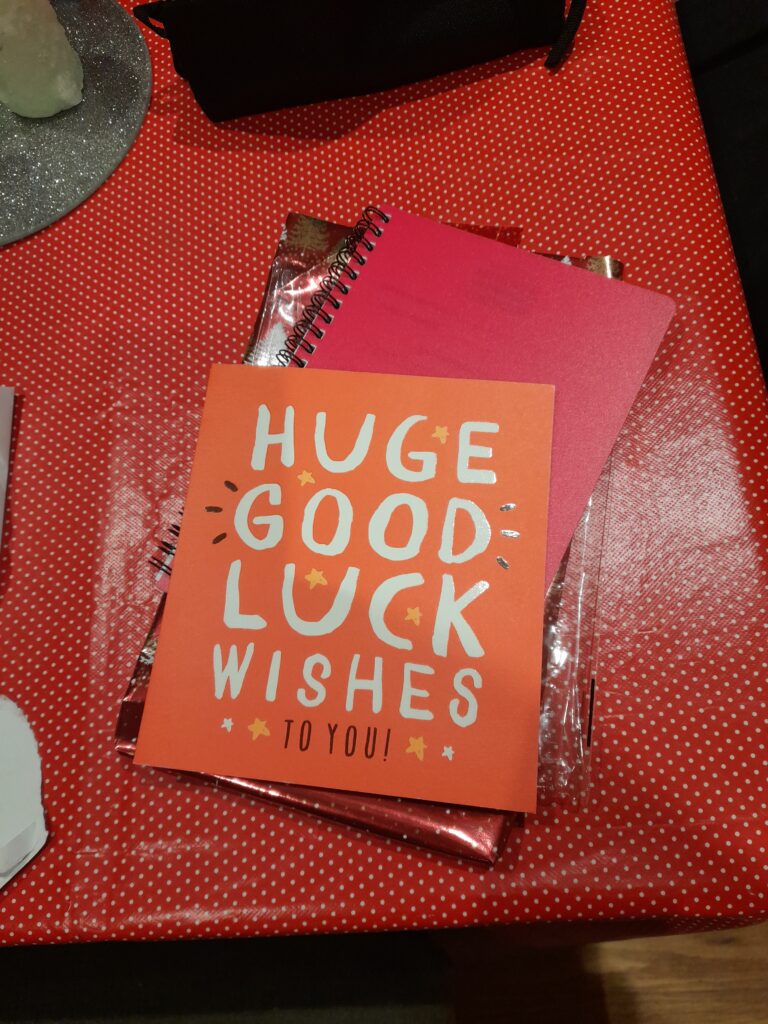 a red card with “huge good luck wishes to you” on it sitting on some note pads on a table covered with a red spitted tablecloth.