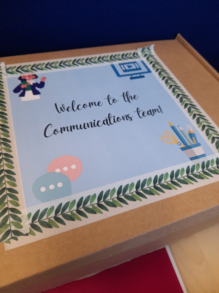 a cardboard box with a paper saying welcome to the communications team on it and images of speech bubbles, a computer, pen pit, and a person.