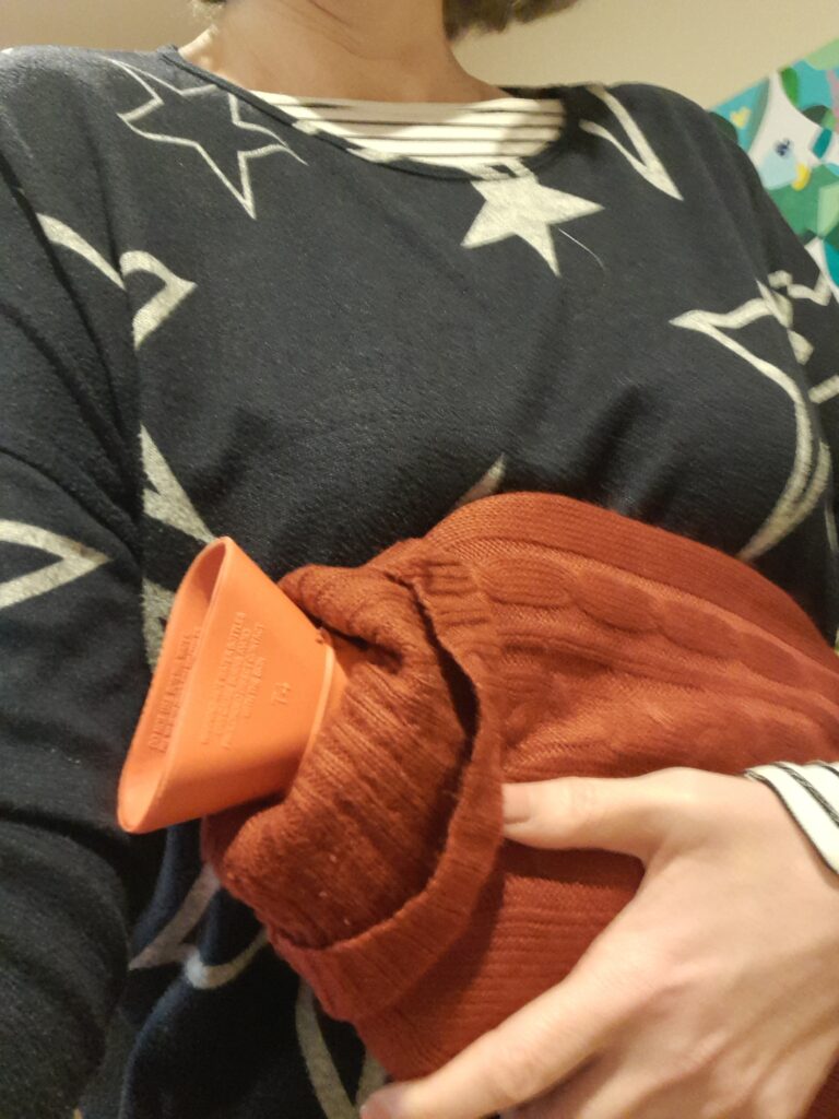 an orange hot water bottle in a burnt orange cover being held to someone’s body.