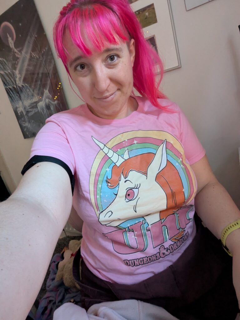 a woman with light skin tone and pink hair wearing a pink t-shirt with a unicorn and a rainbow on it and the words Uni Dungeons and Dragons on it.