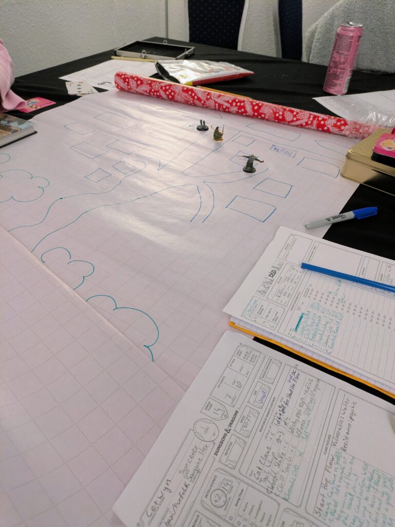 a large table with wrapping paper laid out on it with the back side showing and a very rough hand drawn map on it with some miniatures on it. Around the map are some Dungeons and Dragons character sheets, pens and drinks.