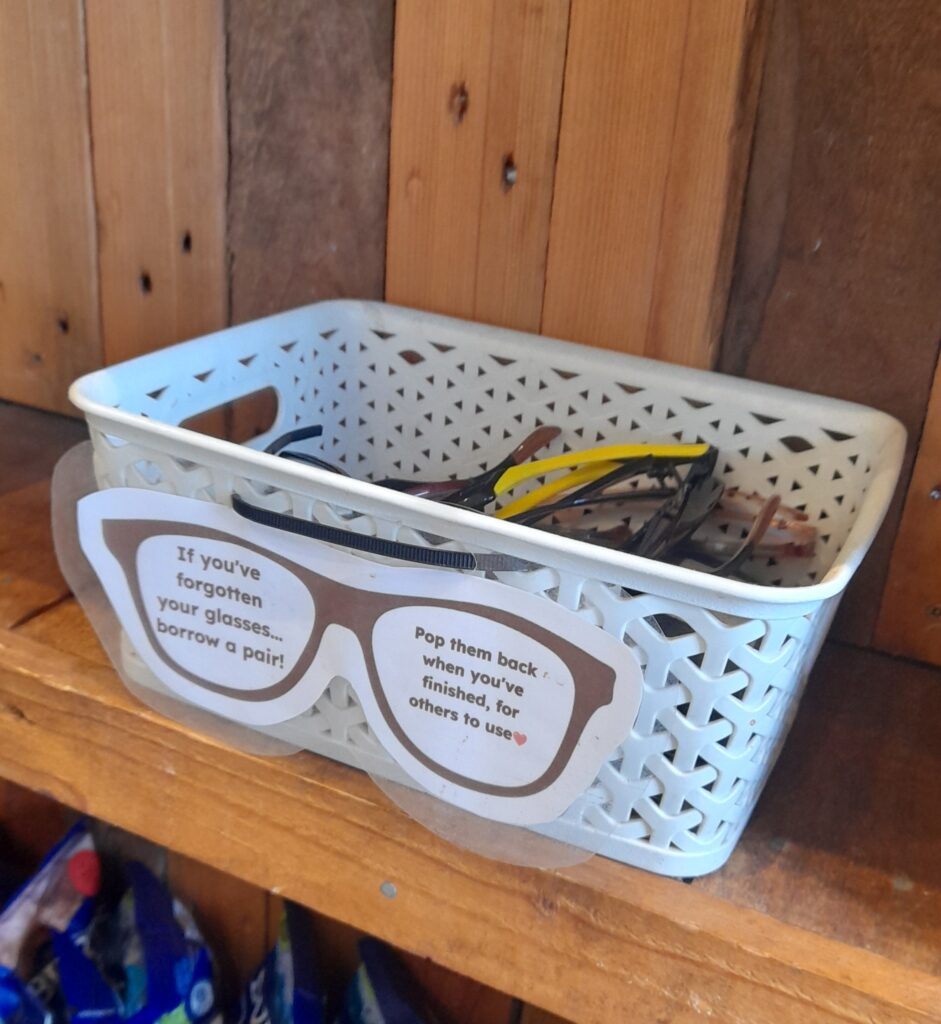 a small basket on a shelf filled with glasses and a sign in the shape of glasses on the front reading if you’ve forgotten your glasses borrow a pair, pop them back in when you’ve finished for others to wear.