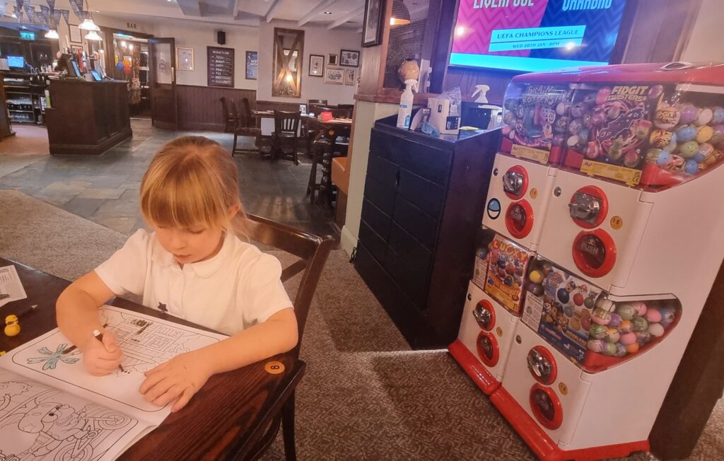 a young girl with long blonde hair, sitting at a table at a pub and colouring in a colouring book. Behind her are four machines filled with balls containing surprises.