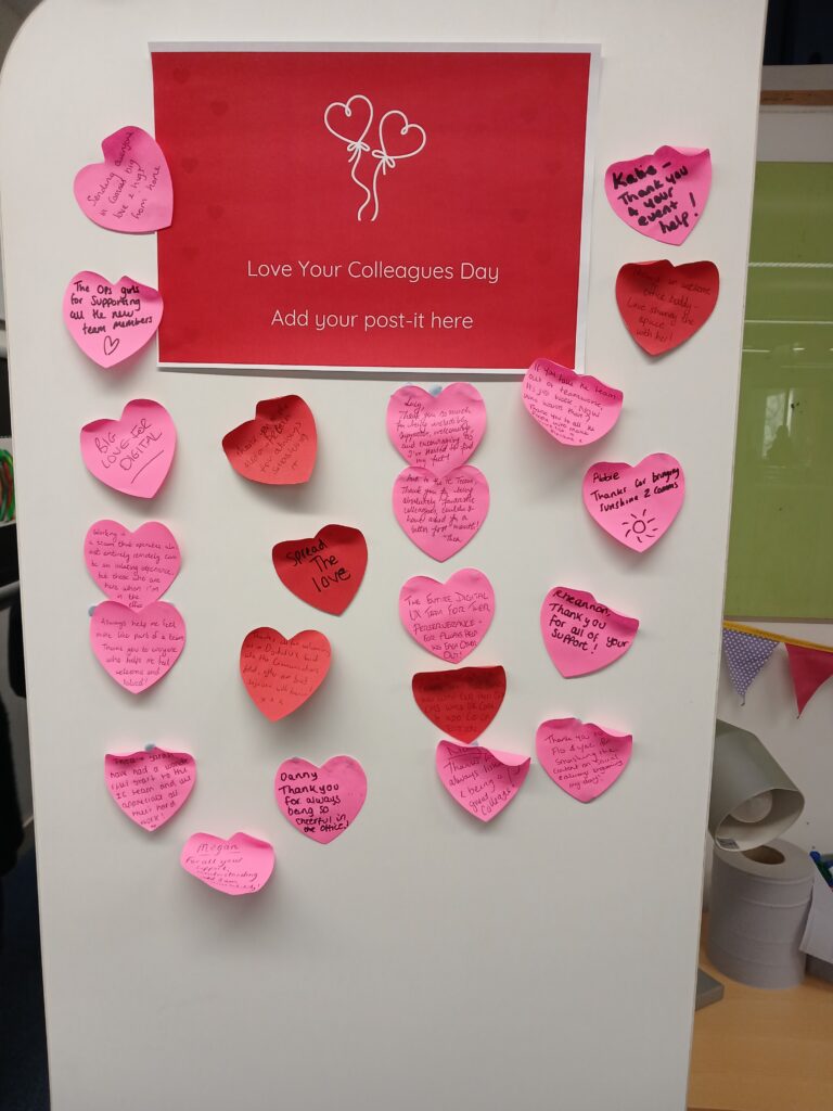A tall white board with red sign on it saying love your colleague day add your post here, with an image of balloon hearts above it. Under and around the sides of the sign are over 20 heart shaped pink and red post its with writing on them.