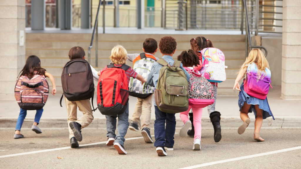 8 primary school aged children in colourful clothes and with colourful backpacks are running away from the camera towards their school building