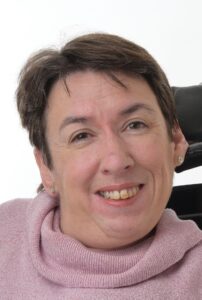 Ruth Meredith, a woman with light skin tone and short brown hair, wearing a pink jumper. She is sitting in a chair with a headrest behind her and smiling at the camera.