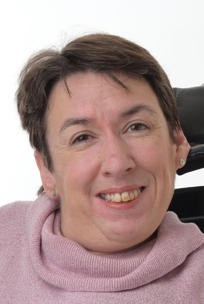 Ruth Meredith, a woman with light skin tone and short brown hair, wearing a pink jumper. She is sitting in a chair with a headrest behind her and smiling at the camera.