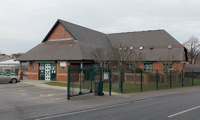 a red brick building which acts as a doctors surgery with car park outside and green fencing around it.