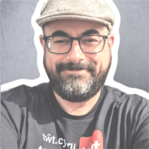 Jaz, a man with light skin tone and short black beard, wearing a toot.wales Tshirt, glasses and flat beige cap.