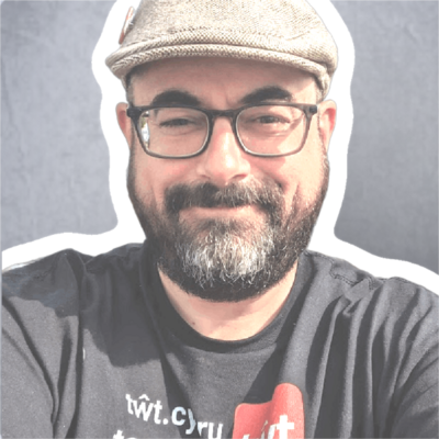 Jaz, a man with light skin tone and short black beard, wearing a toot.wales Tshirt, glasses and flat beige cap.