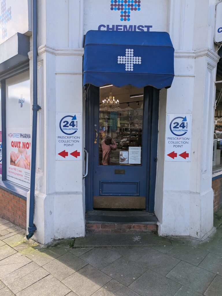 a corner building painted white with a chemist sign above it and a blue door with 24 hour collection point sign either side of the door with arrows pointing left. The door is up a step.