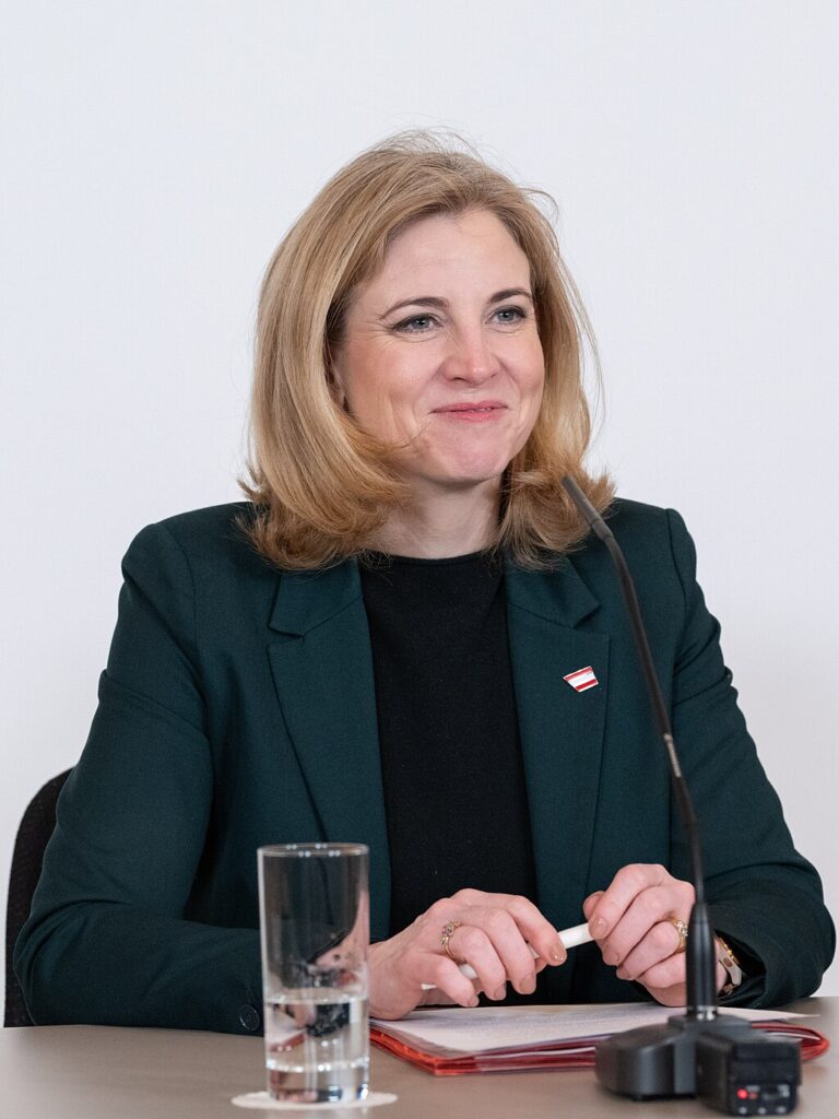 Beate Meinl-Reisinger, a woman with light skin tone and strawberry blonde shoulder length hair, wearing a green suit jacket with black top. She is sitting at a table with a microphone, glass of water, notes and pen in front of her, and smiling.