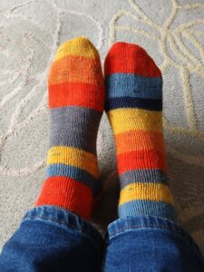 a pair of feet with hand knitted socks on them. The socks are stripey in yellow, orange, red, blue and grey.