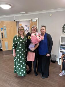 Three women with light skin tone standing together and smiling. One in the middle is holding a large bouquet of flowers, a paper bag and a small purple box.