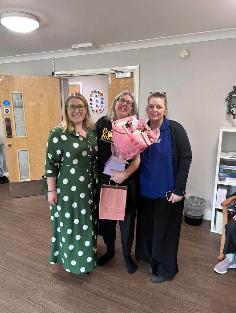 Three women with light skin tone standing together and smiling. One in the middle is holding a large bouquet of flowers, a paper bag and a small purple box.