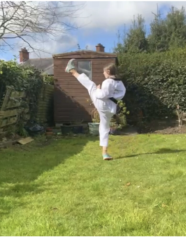 A young woman in a garden with green grass and a blue sky. She is wearing a white martial arts suit and is kicking her left leg above her head