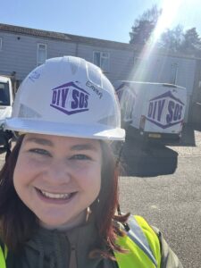 a woman with light skin tone and shoulder length red deep red hair wearing a white hard had with DIY SOS on it and a high vis jacket, stands smiling with a DIY SOS truck and a building behind her.