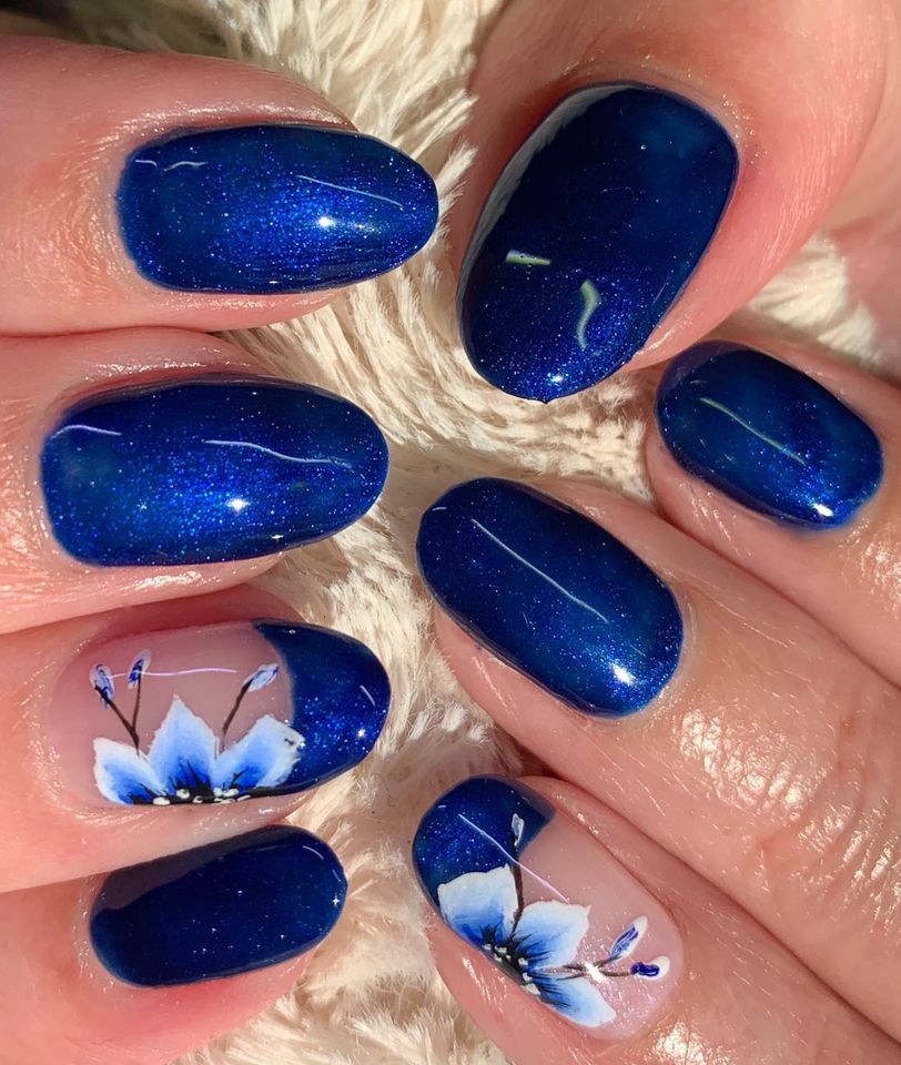 fingers with light skin tone and beautiful sparkly dark blue fake nails, one finger has a solid blue tip and clear paint under it with a delicate white and blue flower over it.