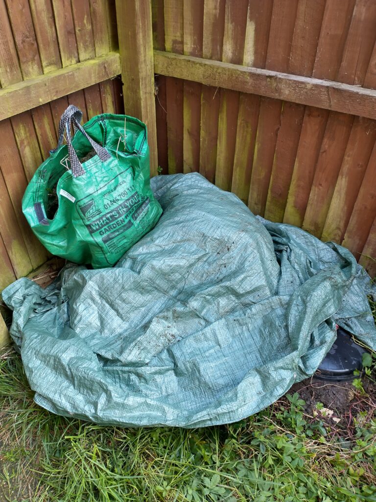 a corner of a garden surrounded by a tall wooden fence. A large tarp covers part of it and on top of that is a green garden waste bag.