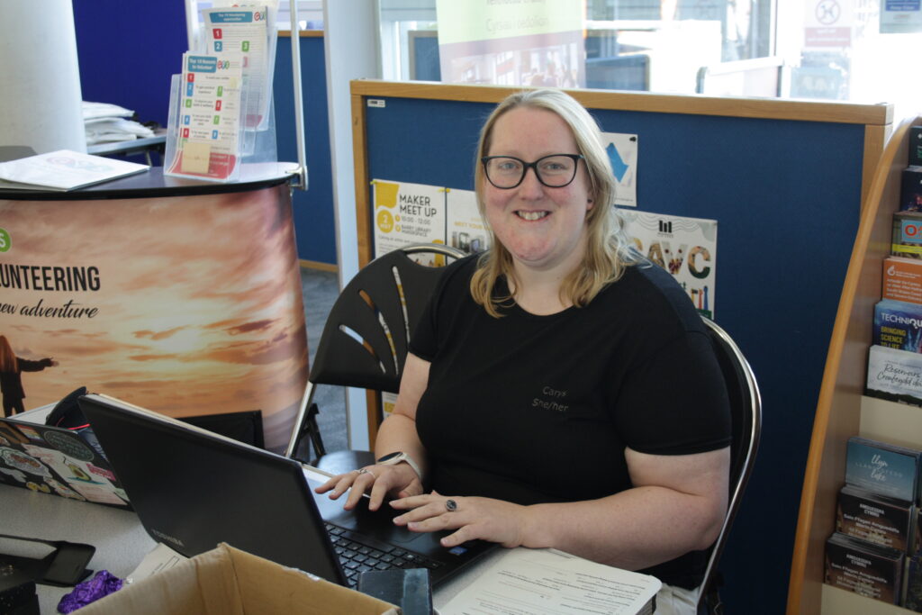 a woman with light skin tone and shoulder length blonde hair wearing a black t-shirt sat at a table typing on a laptop and smiling at the camera.