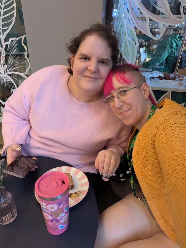 Two women sat at a cafe table. One is tall with light skin tone and short, dark, curly hair, wearing a a light pink jumper. She has beautiful sparkly nails with black cats on them. The other has light skin tone and bright pink hair tied back. She is wearing a yellow knitted cardigan, a very short playsuit and a sunflower lanyard. The are leaning into each other and smiling at the camera. On the table in front of them is a small plate with a slice of banana bread on it and a reusable pink coffee cup decorated in flowers.