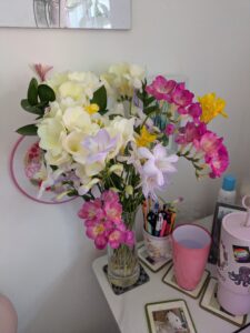 A small bouquet of spring flowers of white, pink, yellow, and lilac, in full bloom in an elegant vase sitting on a desk beside a mug of pens and a pink glass of water.