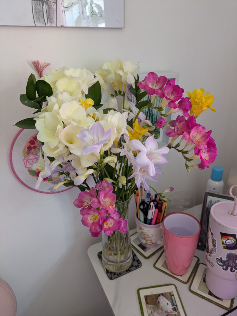 A small bouquet of spring flowers of white, pink, yellow, and lilac, in full bloom in an elegant vase sitting on a desk beside a mug of pens and a pink glass of water.