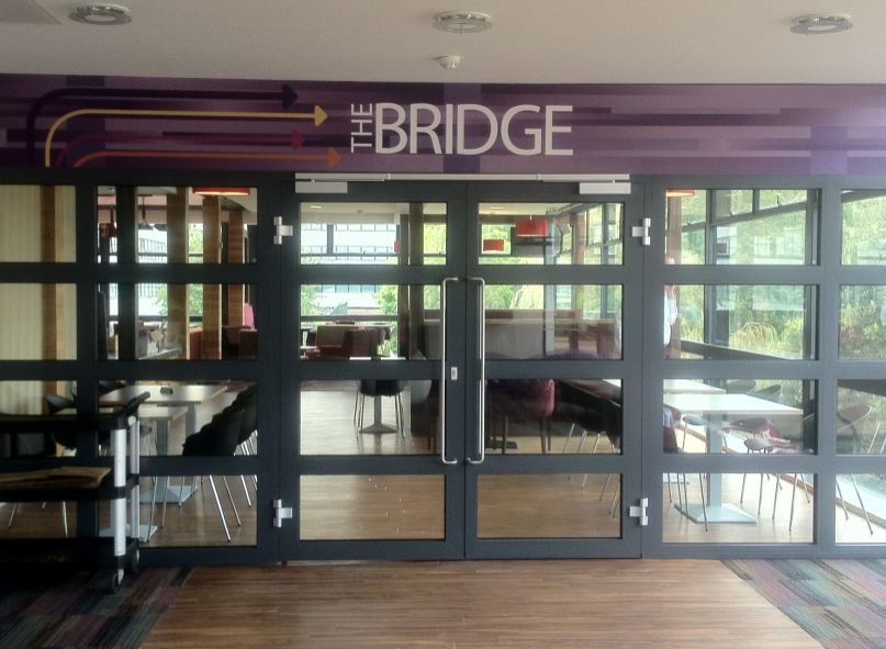 a set of glass panelled doors under a sign reading “The Bridge.” Through the doors seating and tables are visible in a bar.
