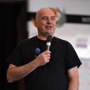 Johannes, a man with light skin tone and bald head standing on stage holding a microphone.